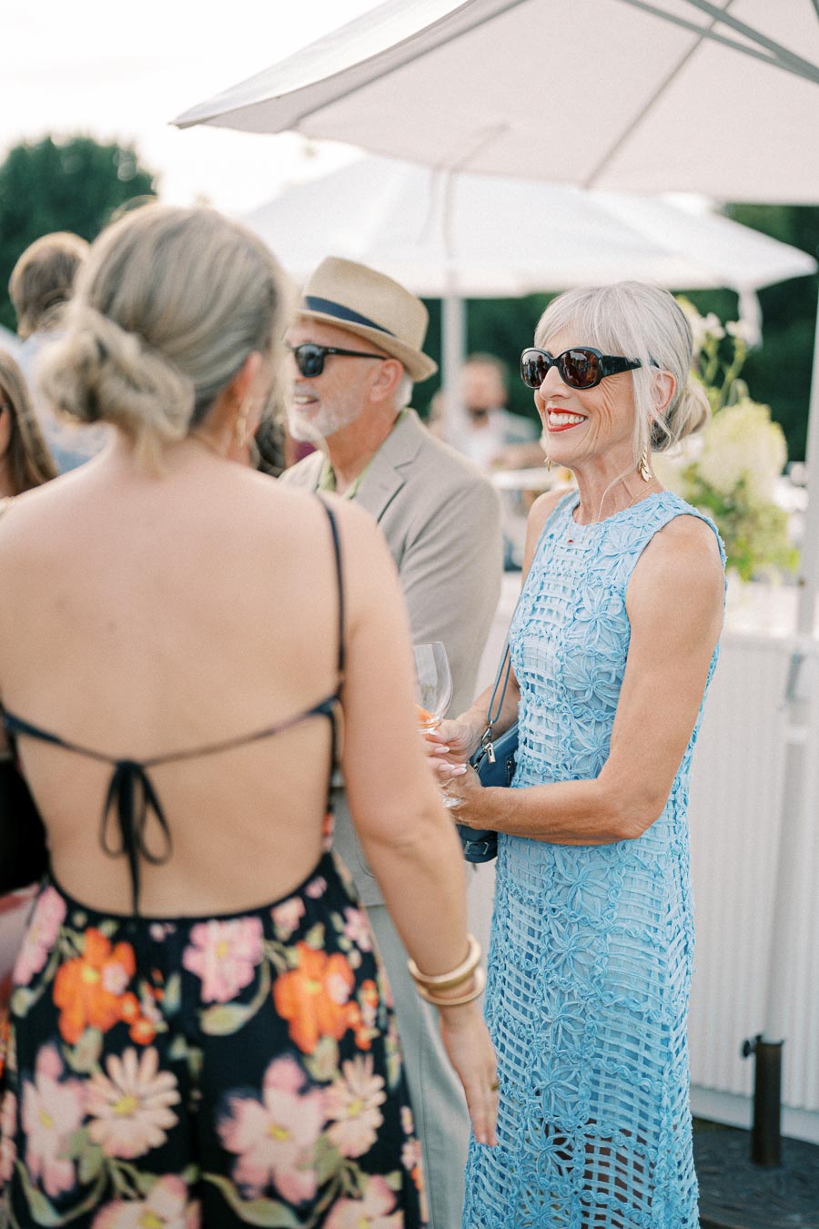 A group of stylishly dressed adults socializing at an outdoor summer event, with a woman in a blue dress and sunglasses smiling under a white umbrella.