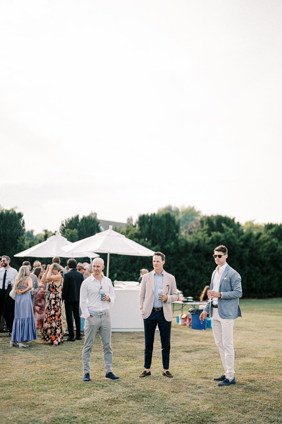 Three men stylishly dressed at an outdoor event, holding drinks. Behind them, guests mingle under white umbrellas, with greenery in the background. The scene is set on a grassy area, capturing a relaxed, social atmosphere on a sunny day.