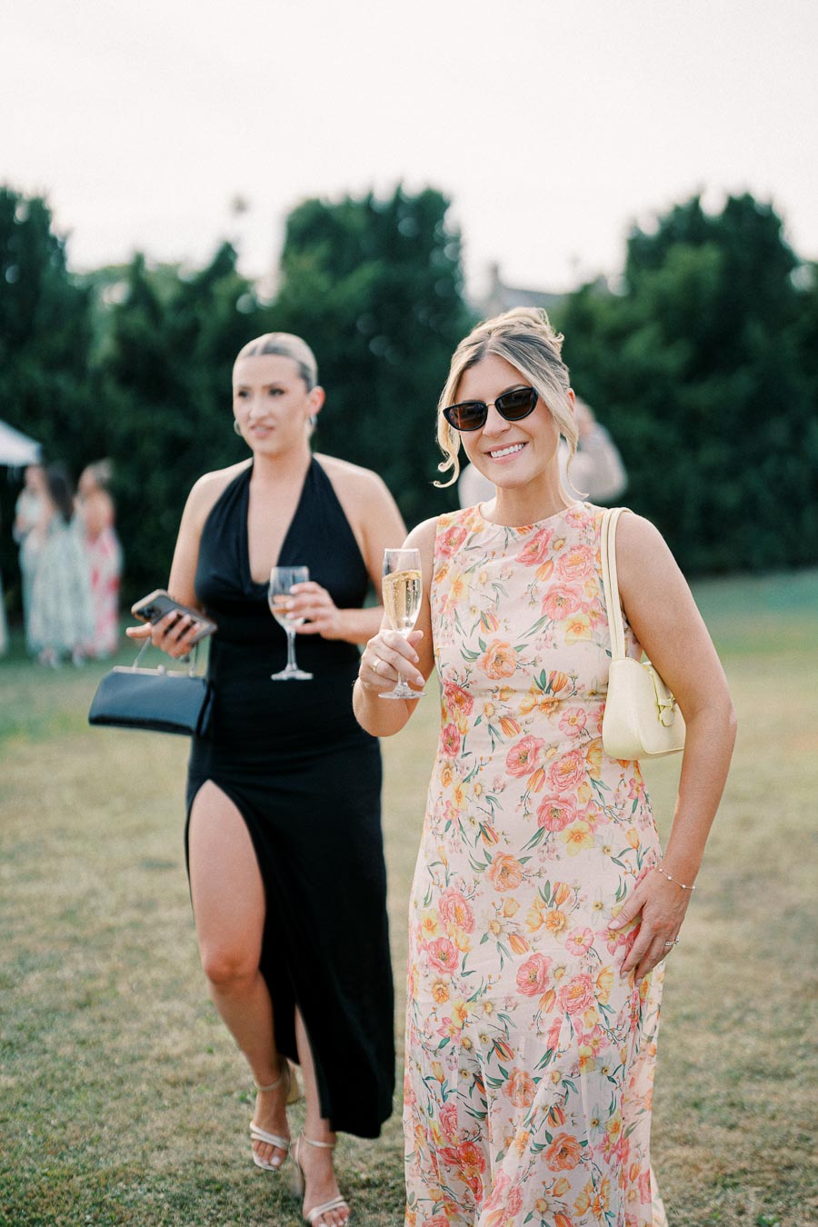Two women enjoying an outdoor event, one in a floral dress with sunglasses and a glass of champagne, and the other in a black dress with a thigh-high slit, holding a glass and a clutch.