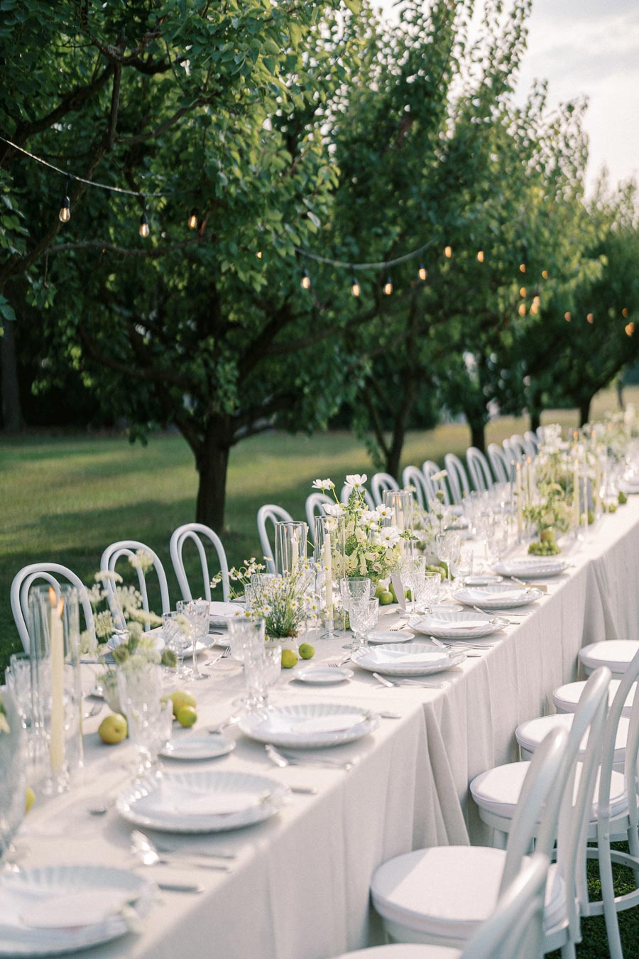 Elegant outdoor wedding reception setup with a long table adorned with white floral centerpieces and green accents, surrounded by white chairs under string lights in a lush garden setting.