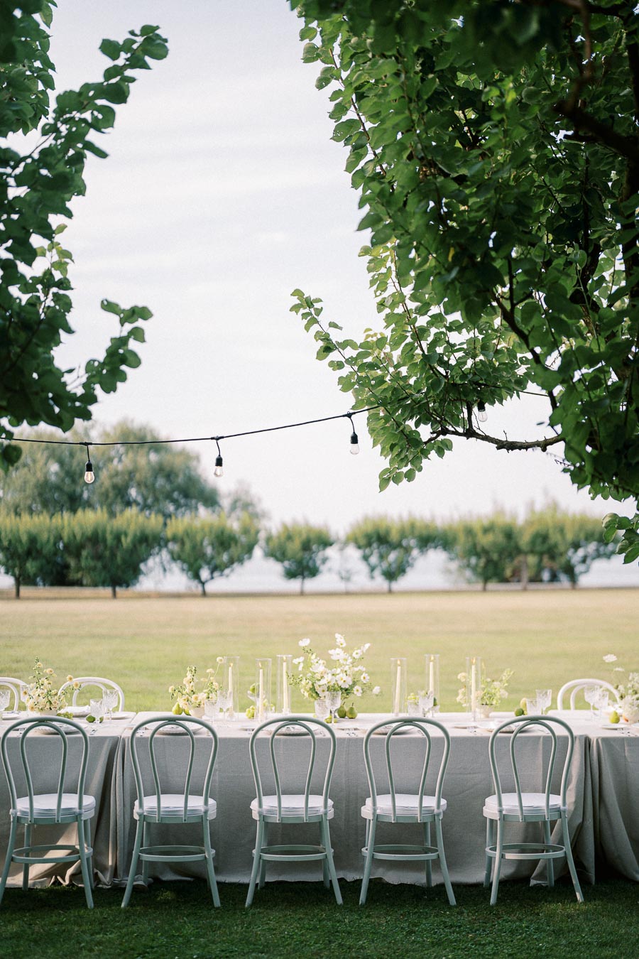 Outdoor garden wedding reception setup with elegant white chairs and a beautifully decorated table in a lush grassy field, framed by leafy trees and hanging string lights.