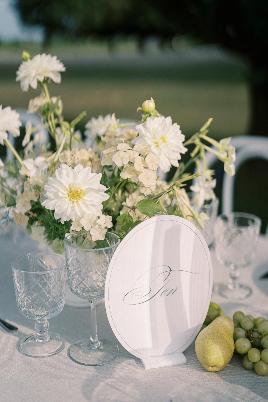 Elegant outdoor table setting with white flowers, crystal glassware, and a personalized table number card. Features fresh pears and grapes, enhancing a sophisticated and natural ambiance.