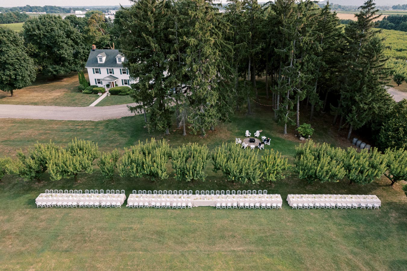 Aerial view of a picturesque outdoor wedding setup featuring long tables with white chairs on a green lawn, lush trees, and a charming house in the background. Ideal for weddings and events in a natural setting.