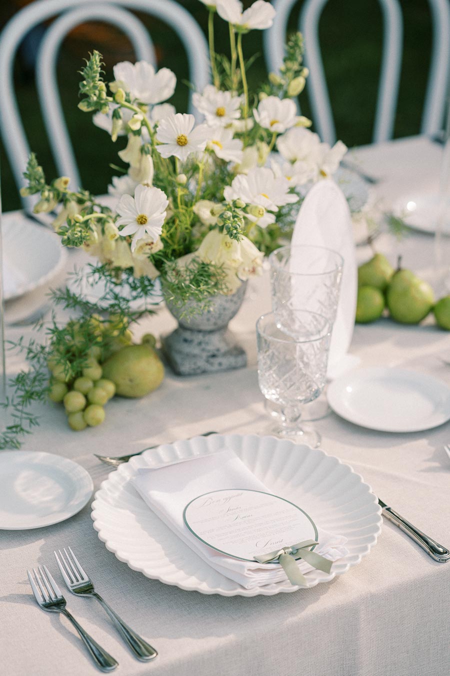 Elegant outdoor table setting with a white floral centerpiece, featuring cosmos flowers in a stone vase. The table is adorned with white plates, crystal glasses, and silver cutlery on a linen tablecloth. Fresh green grapes and pears complement the light and airy aesthetic, creating a sophisticated atmosphere for a garden event or wedding reception.