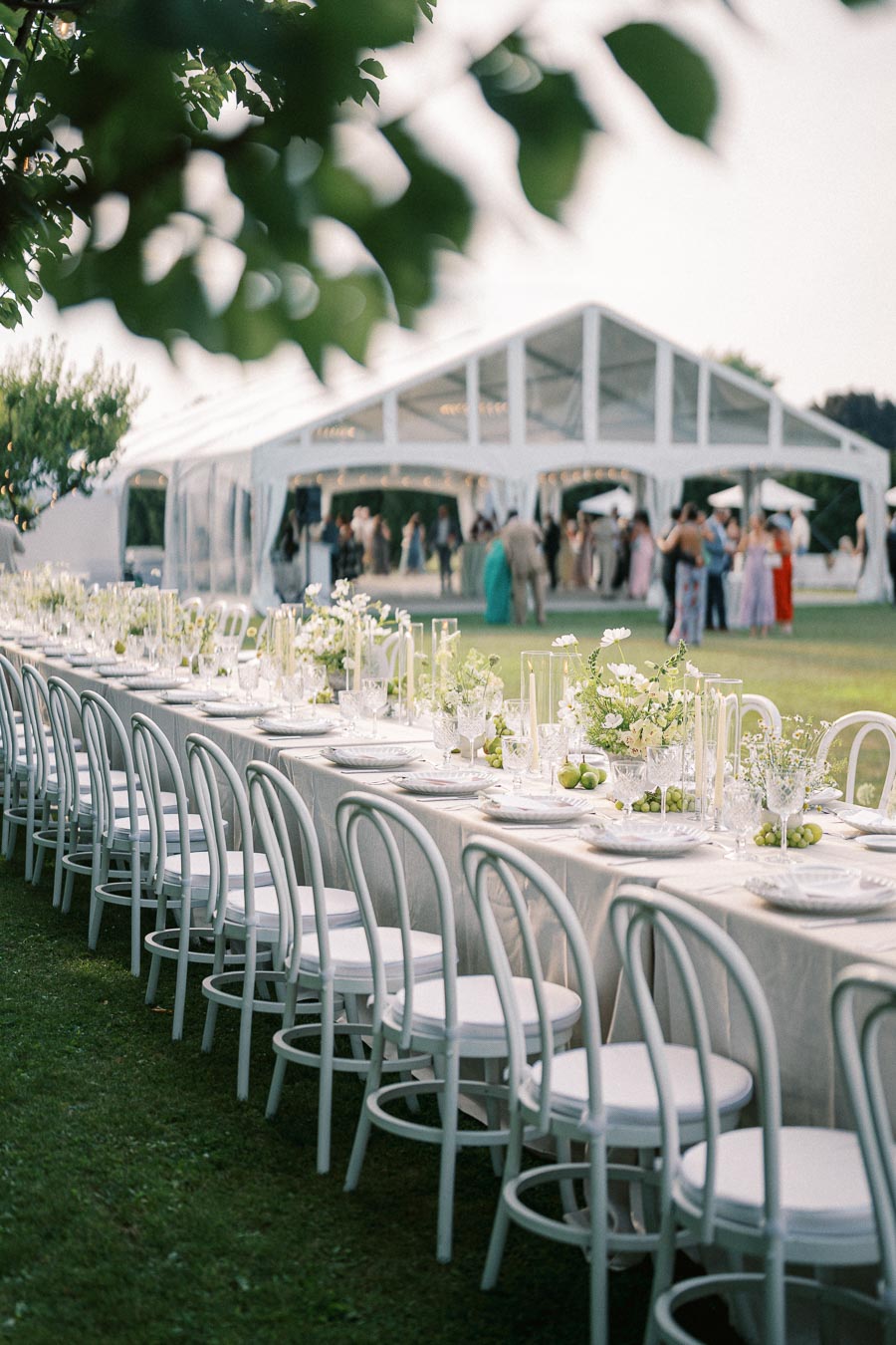 Outdoor wedding reception setup featuring a long, elegantly decorated table with white chairs on a grassy lawn, surrounded by lush greenery, with a clear tent in the background hosting guests.