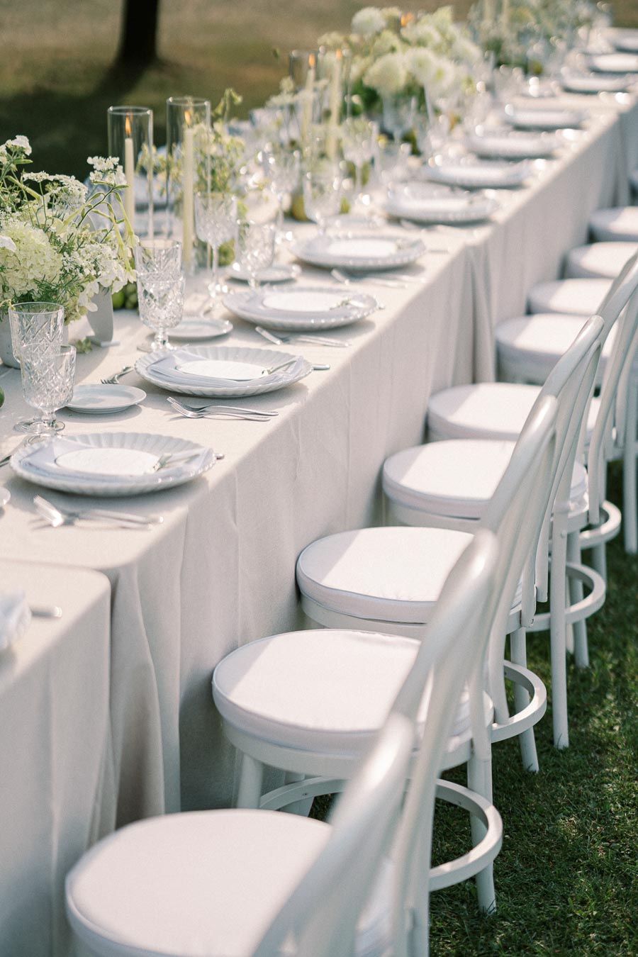Elegant outdoor wedding reception table setting with white chairs, floral centerpieces, crystal glassware, and lit candles, creating a sophisticated and romantic atmosphere.