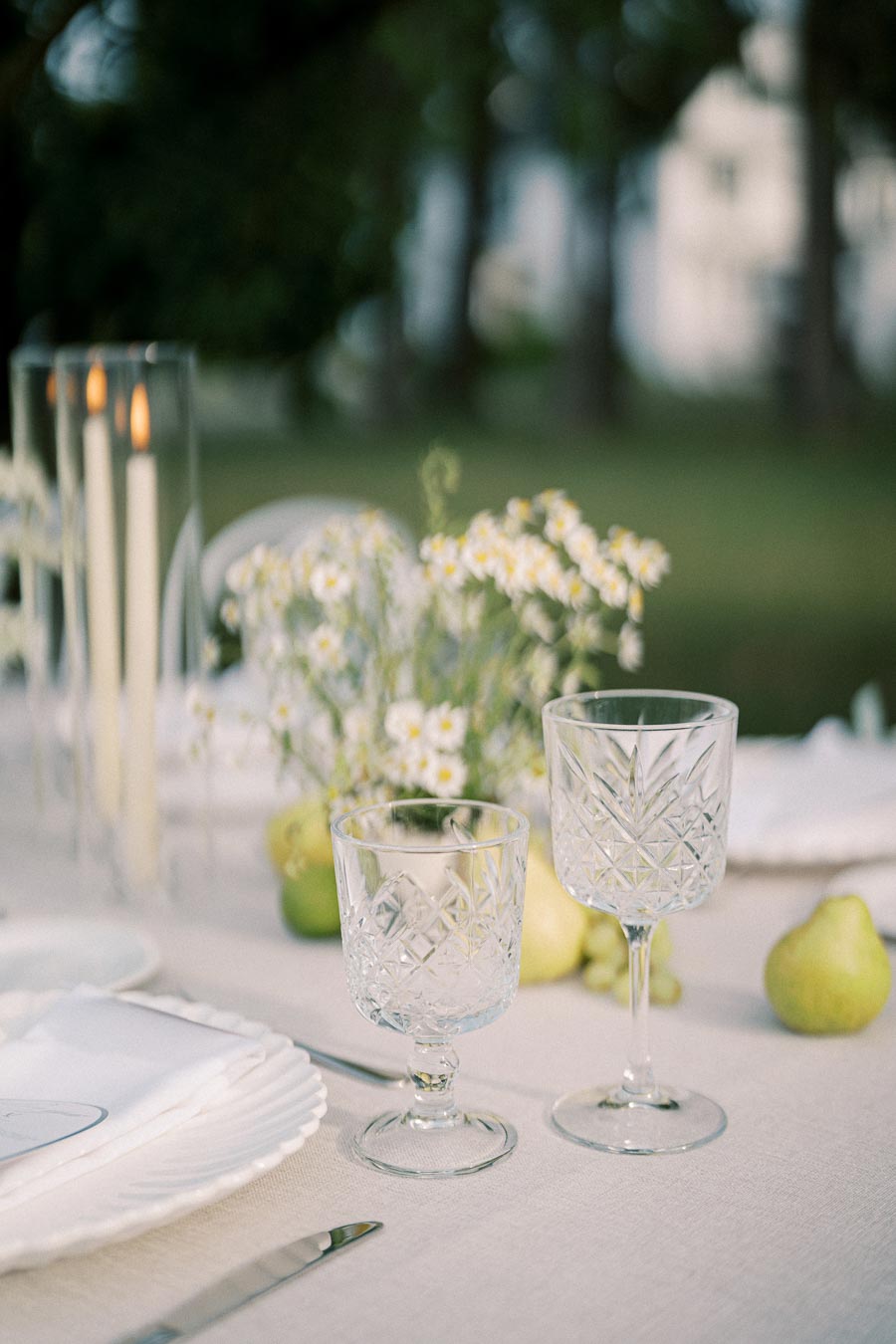 Elegant outdoor dining setup with crystal glassware, white flowers, and candles on a linen tablecloth.