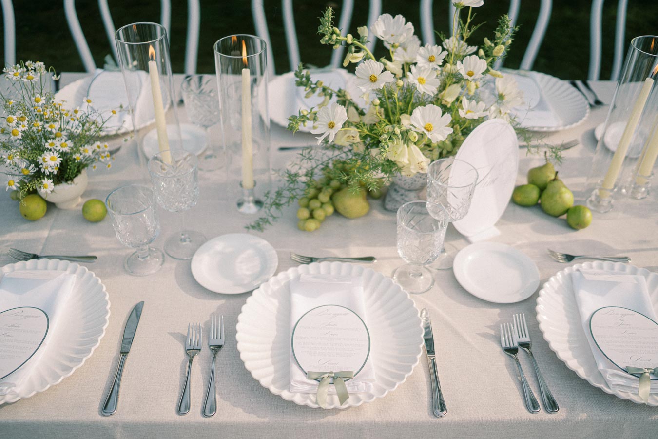 Elegant outdoor wedding table setting with white plates and glassware, adorned with a floral centerpiece featuring white daisies and greenery, surrounded by candles and fresh green pears.