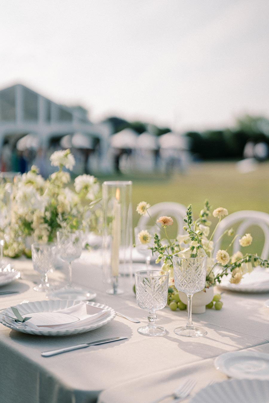 Elegant outdoor wedding reception table setup with crystal glassware, white floral centerpieces, and soft linen tablecloth under a sunny sky.