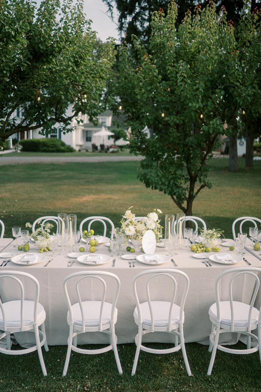 Elegant outdoor dining setup in a garden setting featuring a long table with a white tablecloth, surrounded by white chairs. The table is adorned with white floral arrangements, candles, and fresh green pears, set under lush green trees. Perfect for a wedding or formal event.