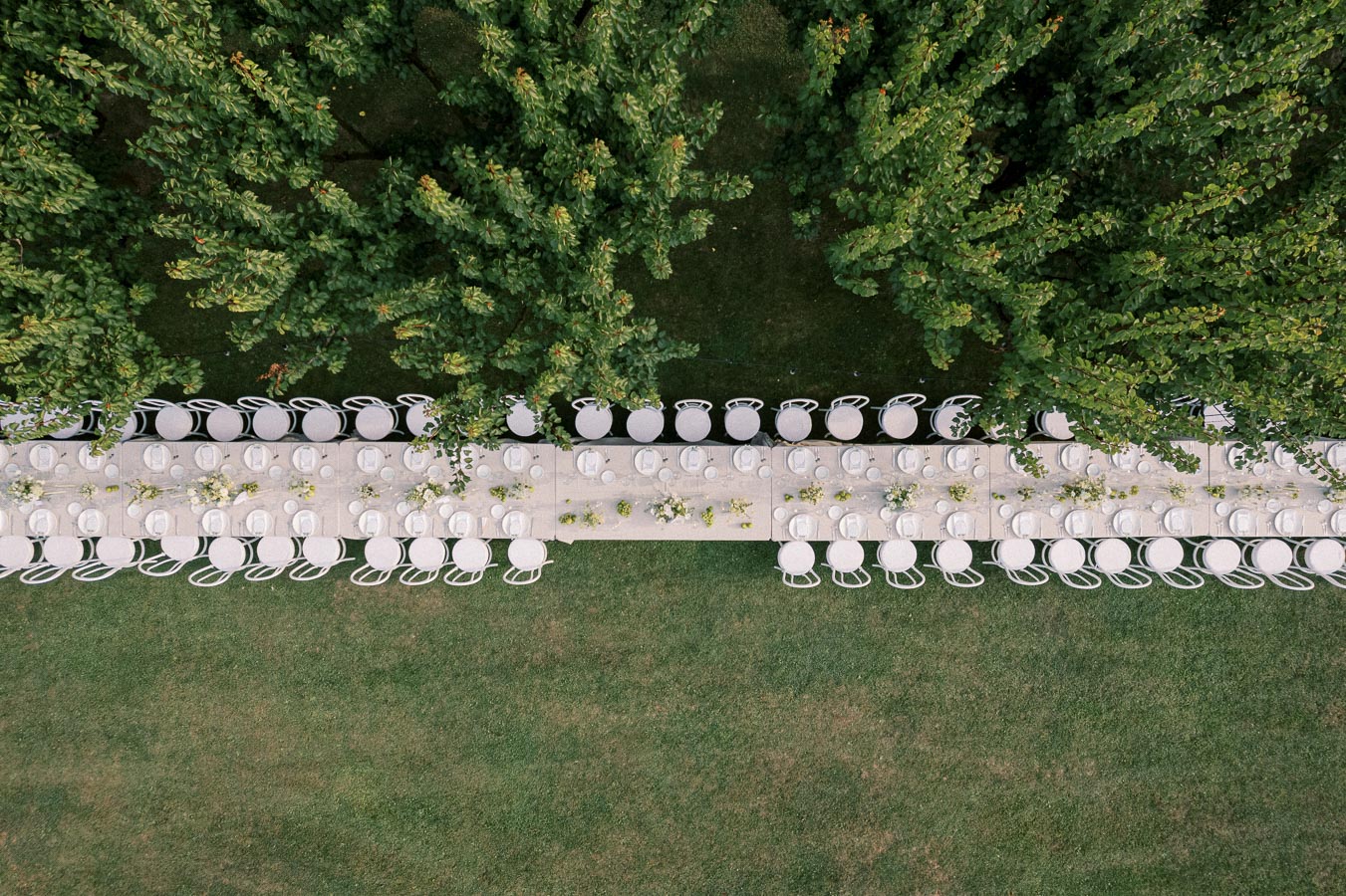 Aerial view of a long, elegantly set outdoor dining table surrounded by white chairs on a grassy lawn, with lush green trees providing a natural border, ideal for a garden wedding or outdoor event.