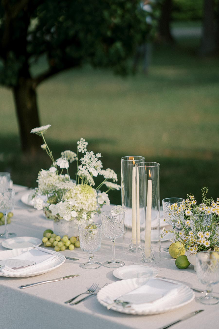 Elegant outdoor dining table setting with white floral arrangements and lit candles in glass holders. Crystal glassware and white plates are adorned with fresh green grapes and limes, creating a sophisticated atmosphere in a lush garden setting.