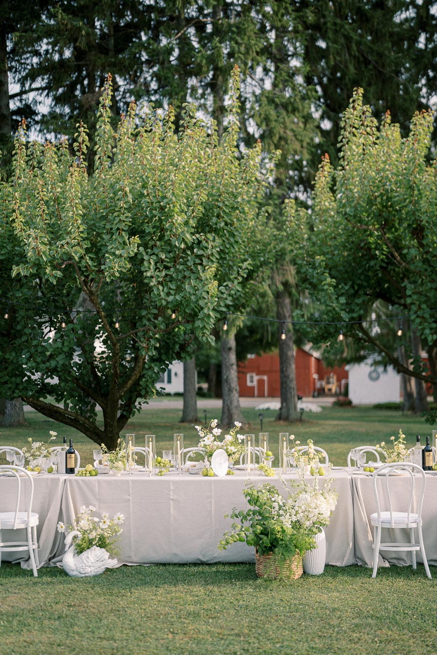 Elegant outdoor wedding reception table setup under lush green trees, featuring white chairs, a beige tablecloth, floral centerpieces, and candles, creating a romantic garden ambiance.