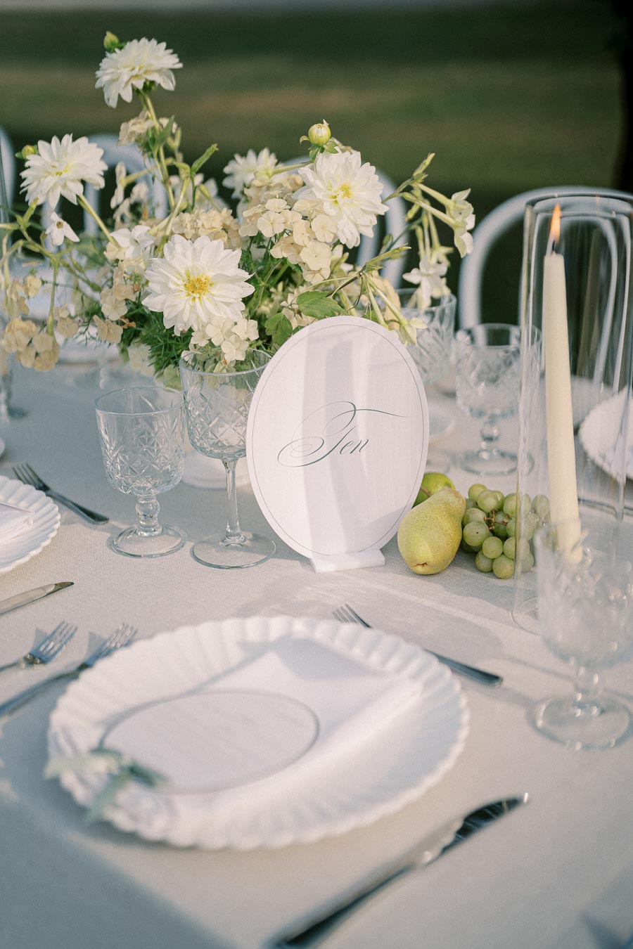 Elegant outdoor wedding table setting with white floral centerpiece, glassware, and fruits, featuring a “Table Ten” sign on a light tablecloth.