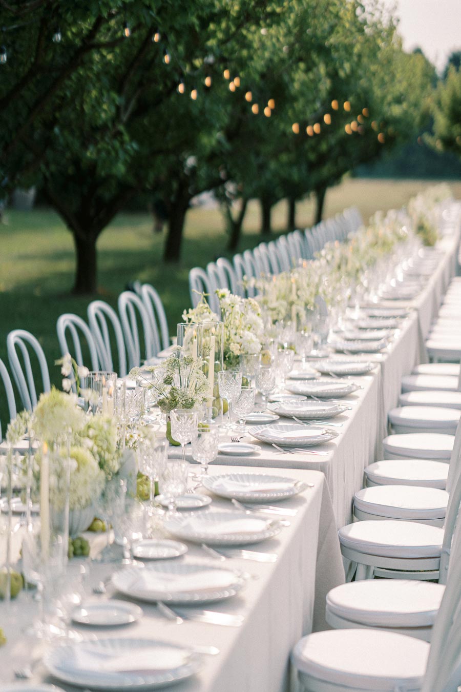 Elegant outdoor wedding reception table setting with white chairs, delicate floral arrangements, candles, and decorative string lights under lush green trees.