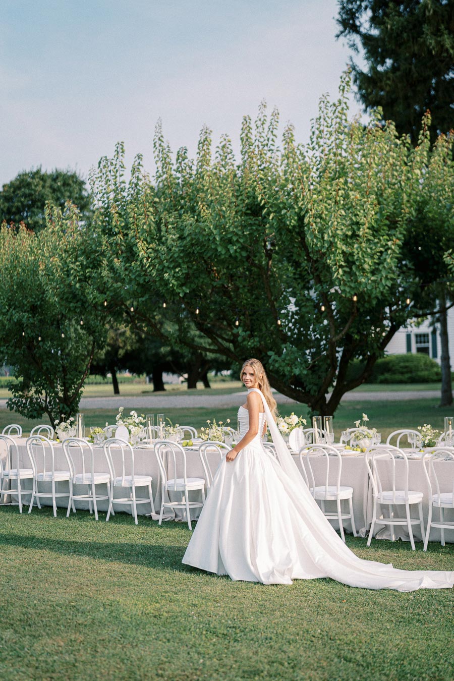 A bride in an elegant white gown stands near a beautifully arranged outdoor wedding reception table, set amidst lush green trees and a clear blue sky, creating a picturesque garden wedding scene.