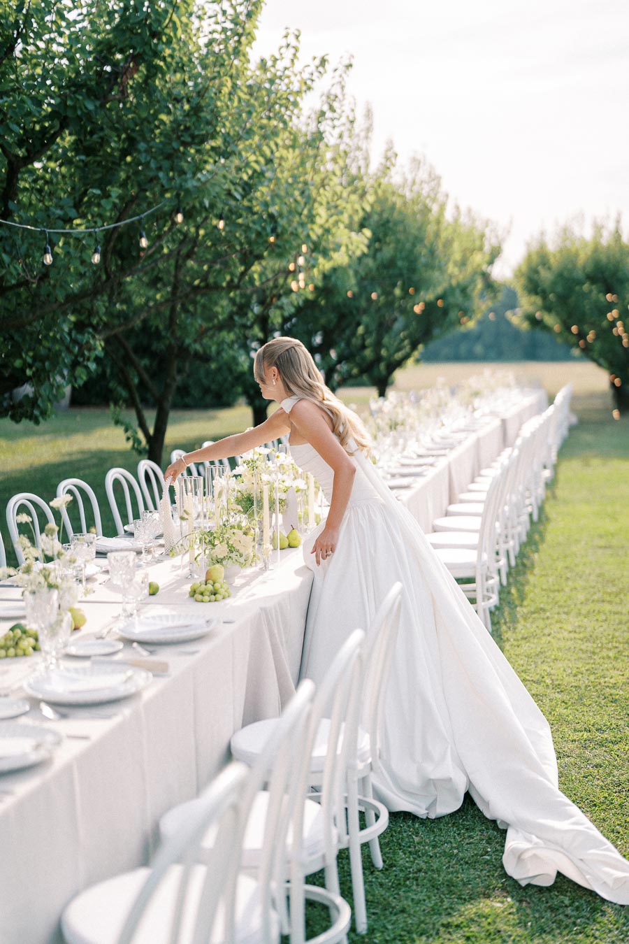 A bride in a white gown arranging a long outdoor wedding table adorned with white linens, elegant glassware, and green floral arrangements under lush trees with string lights.