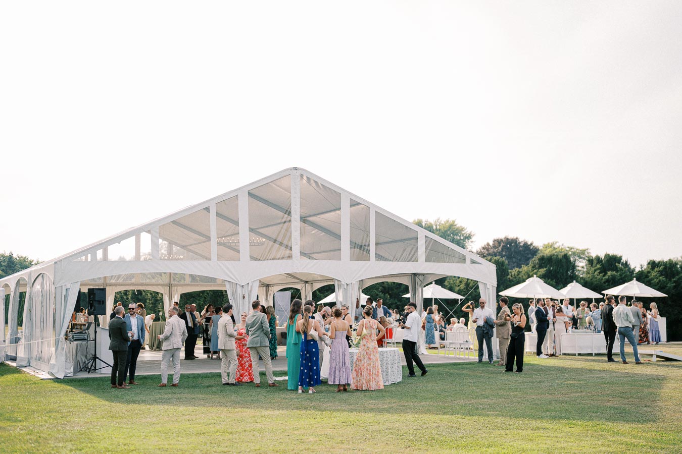 Outdoor wedding reception with guests mingling under a large white marquee tent on a sunny day.