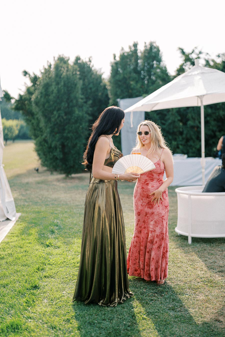 Two elegantly dressed women in conversation at an outdoor event, one in a green gown holding a fan, the other in a red floral dress, standing on a grassy lawn under sunny skies near a white umbrella.