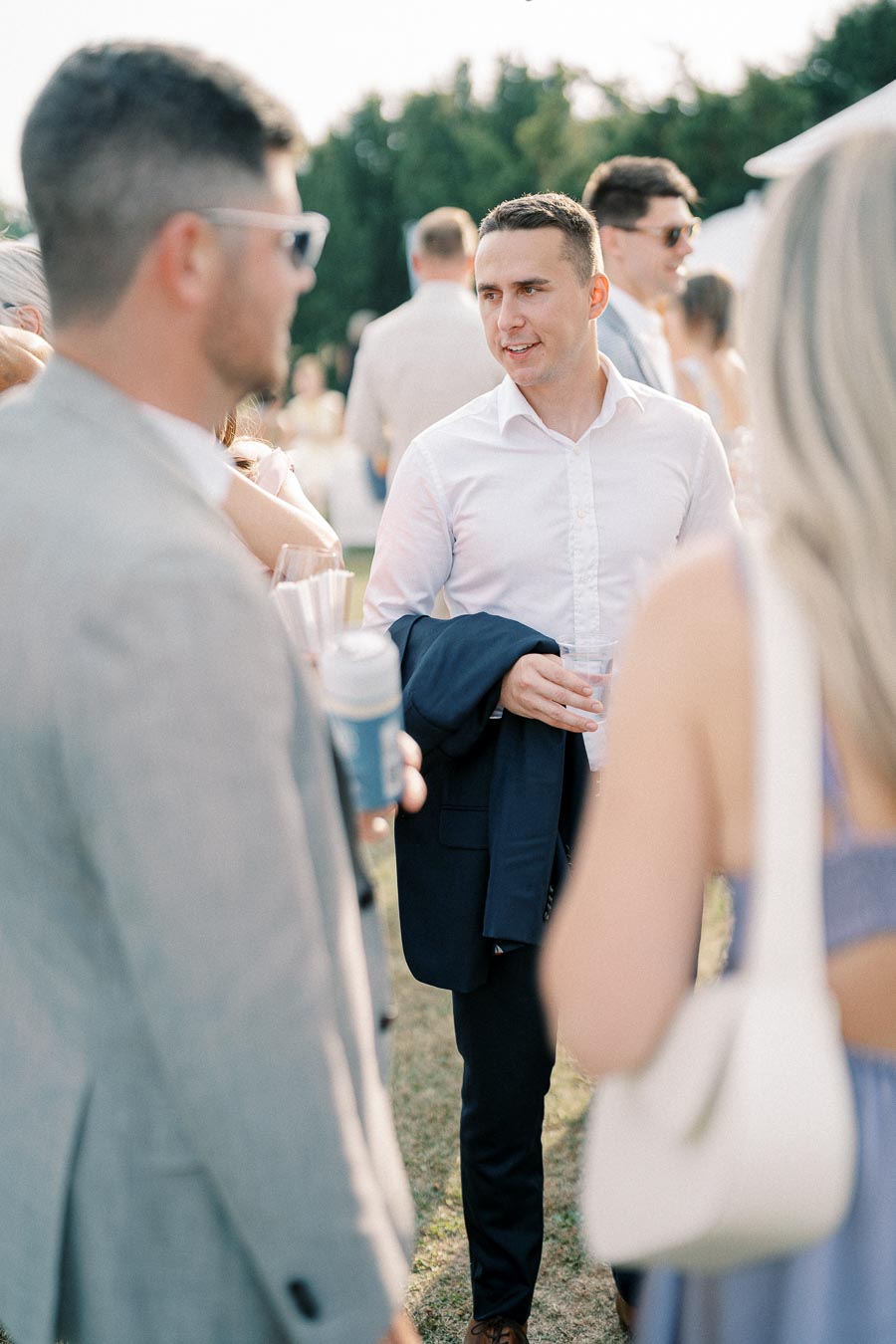 Group of people at an outdoor social event, with a man in a white shirt holding a jacket and drink, engaged in conversation.
