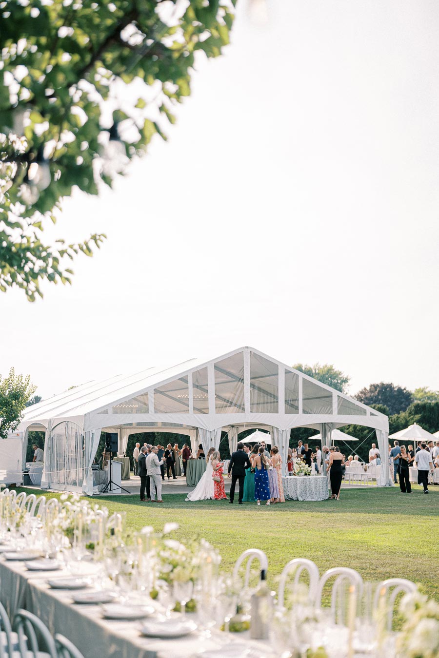 Elegant outdoor wedding reception under a large white tent, with guests socializing on a sunny day, surrounded by green lawn and beautifully set tables adorned with white flowers and glassware.