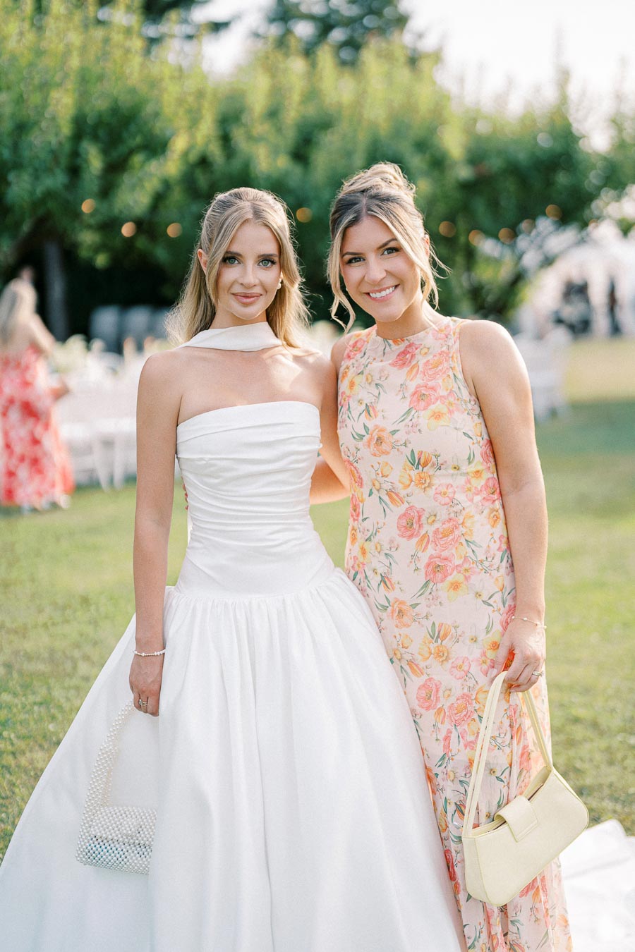 Two women smiling in elegant dresses at an outdoor event, one in a white gown and the other in a floral dress, standing on a grassy field with blurred trees in the background.