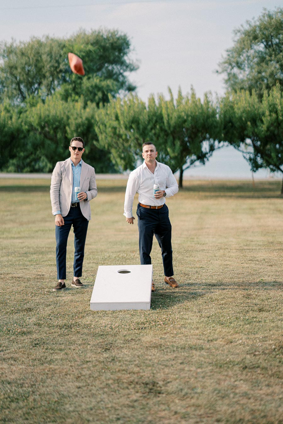 Two men playing cornhole in a grassy field, each holding drinks, with one tossing a bean bag towards the target under a clear sky.