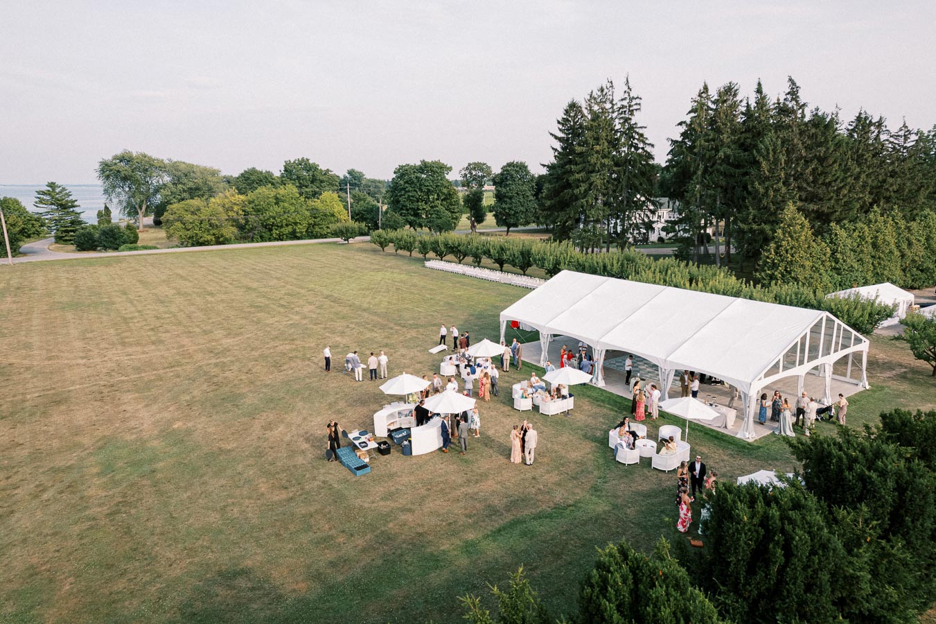 Aerial view of an outdoor event with people gathering near a large white tent on a grassy field, surrounded by trees and clear skies.