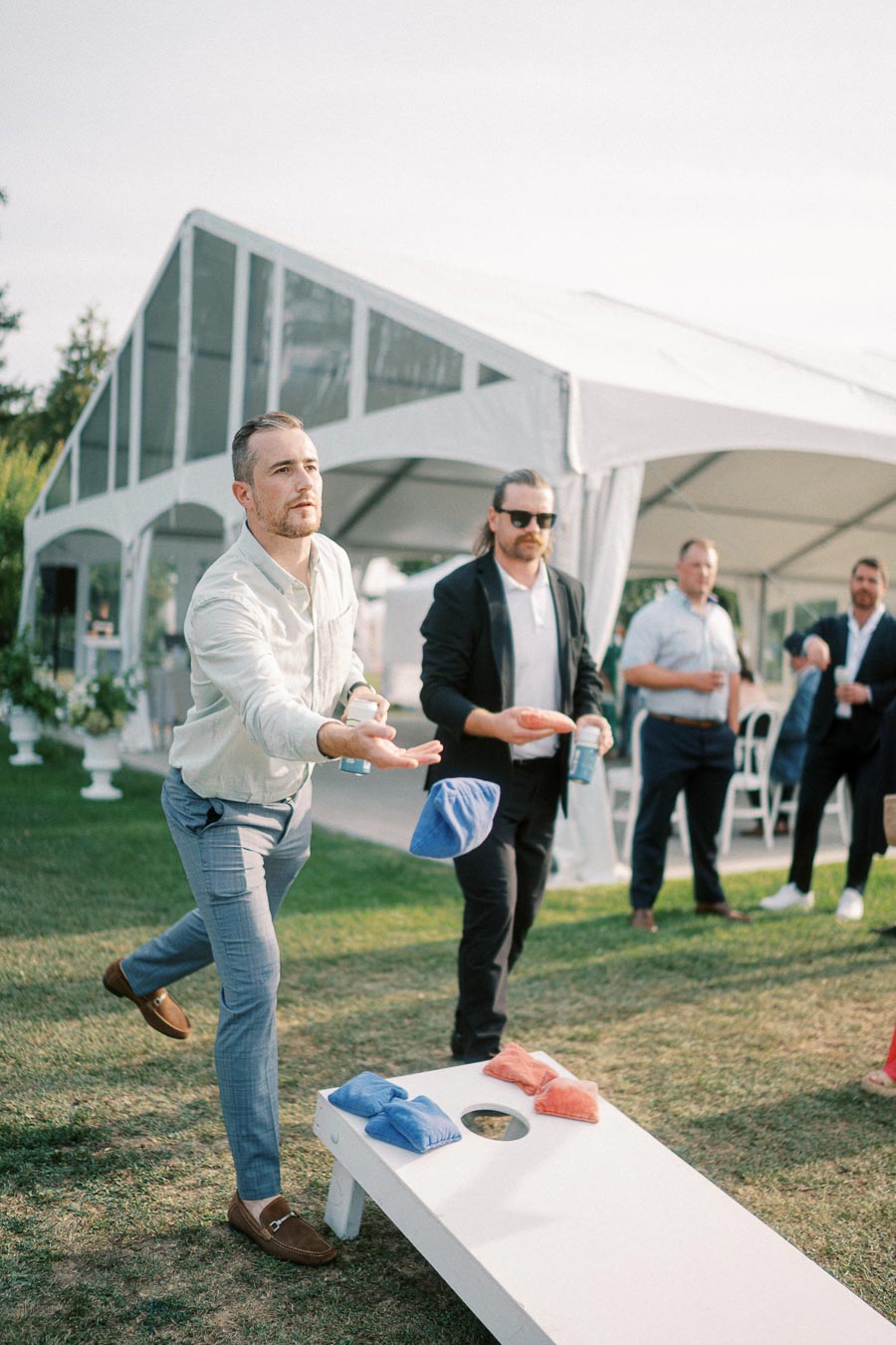 Group of men playing cornhole outdoors near a large white tent during a casual social event, with one man tossing a bean bag as others watch.
