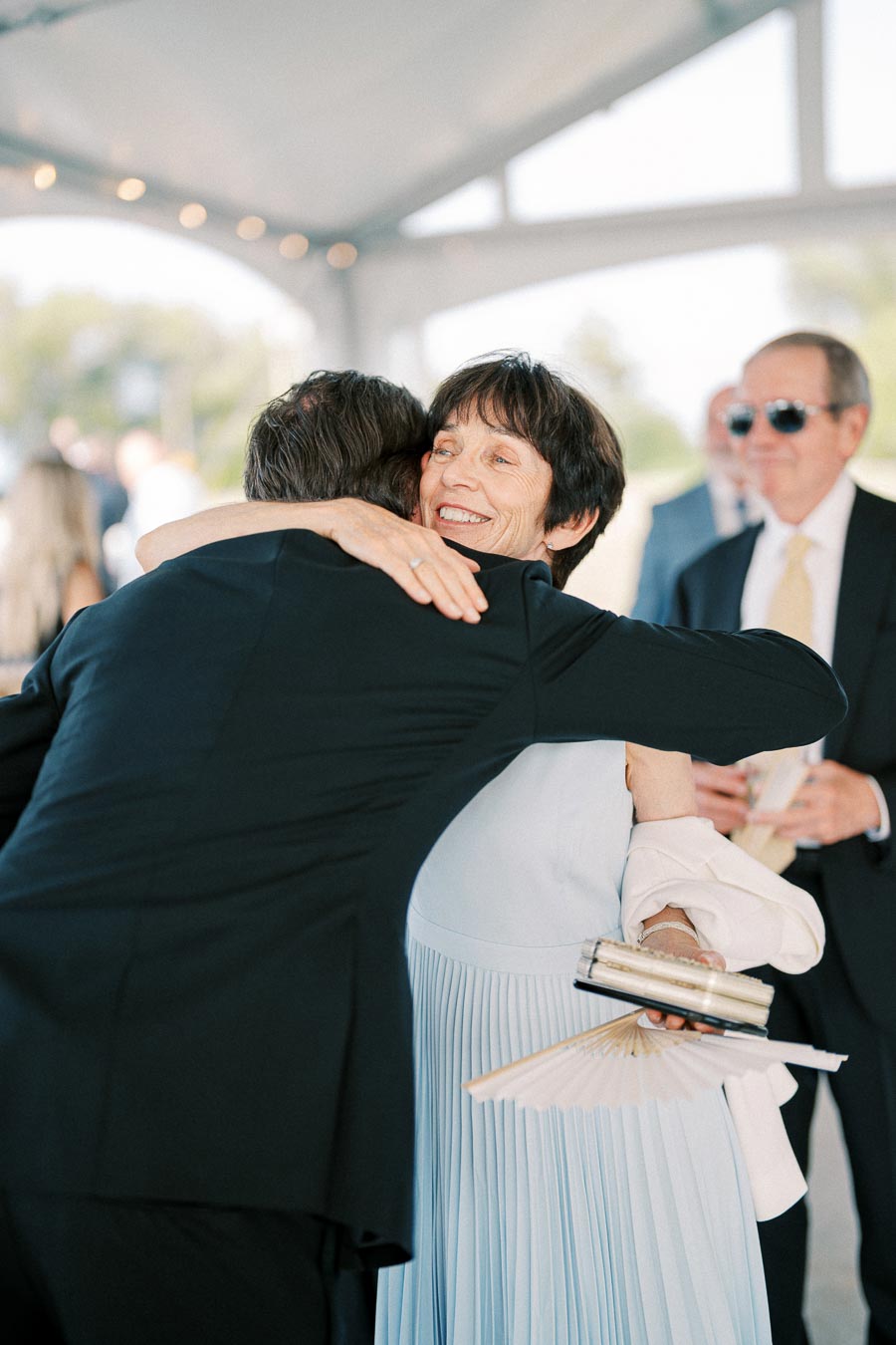Guests sharing a warm embrace at an elegant outdoor event, with a woman in a light blue dress smiling while holding a folding fan.