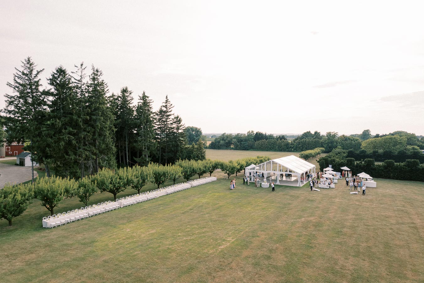 Aerial view of an elegant outdoor event setup in a spacious green field, featuring a large white marquee tent surrounded by neatly arranged tables and chairs, with guests socializing under clear skies amidst lush trees. Ideal for upscale garden weddings or luxury celebrations.