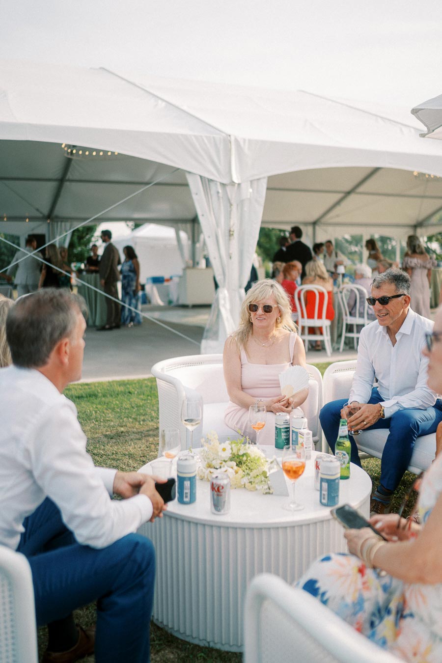 Group of people socializing under a white outdoor tent, seated around a table with drinks and flowers, during a daytime gathering event.