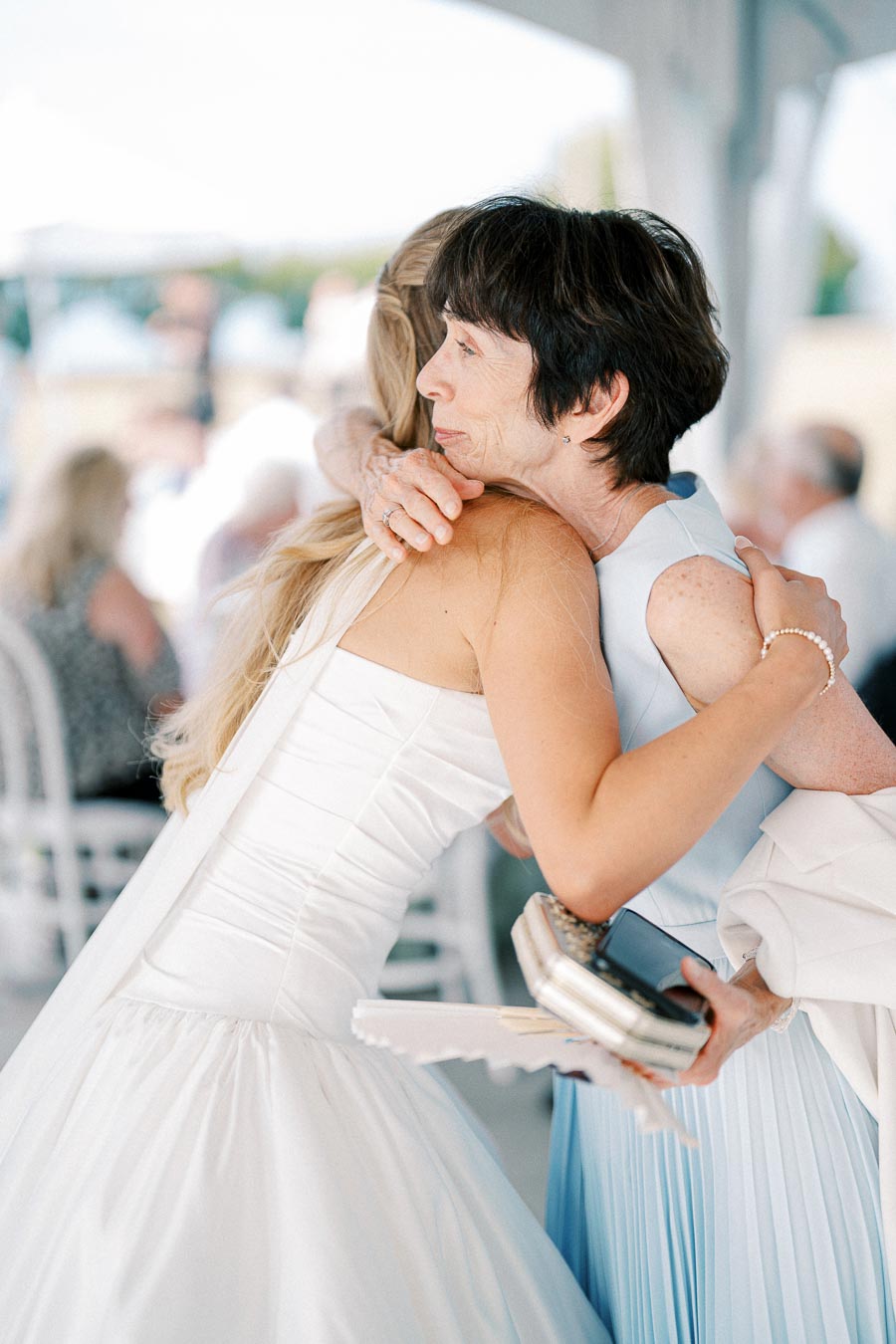 A bride in a white wedding dress warmly embraces a woman in a pastel blue outfit, holding a clutch and phone, during a wedding reception, highlighting a heartfelt moment of connection and emotion.