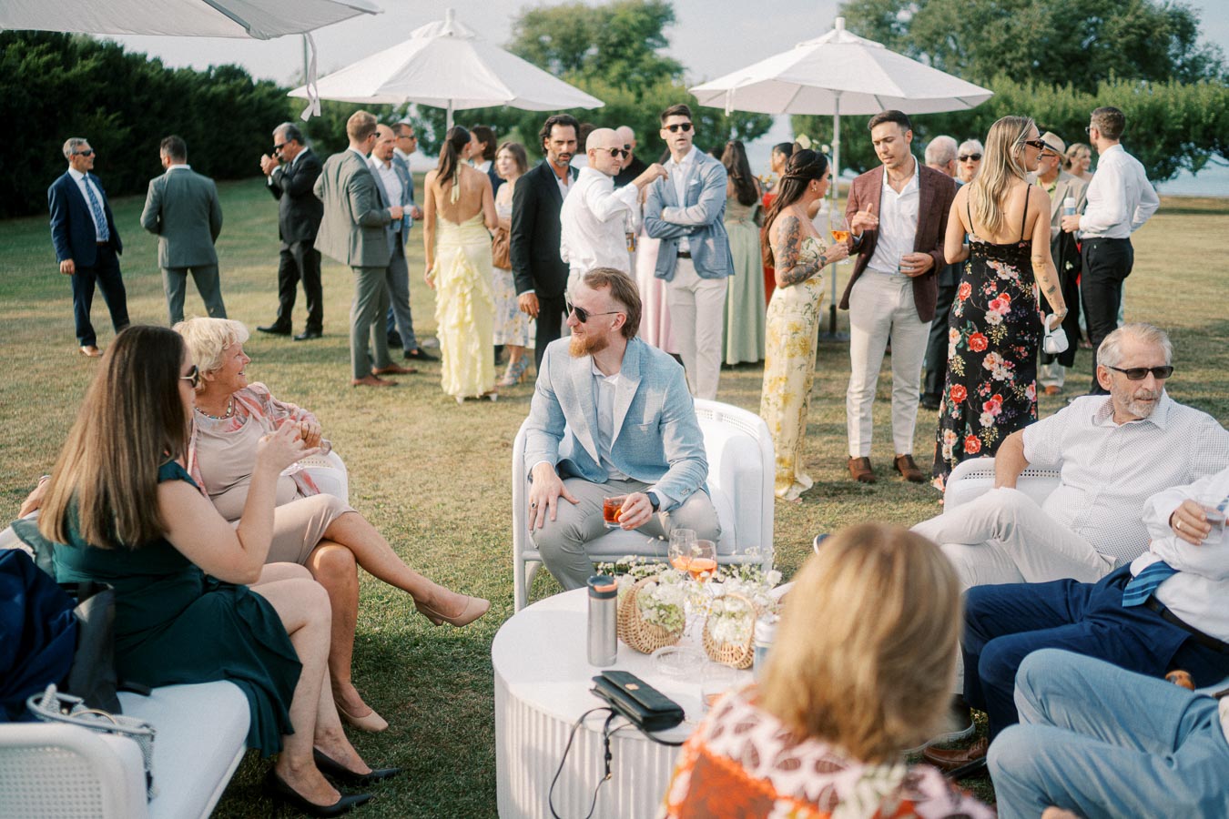 A group of elegantly dressed people at an outdoor event, socializing under white umbrellas on a sunny day.