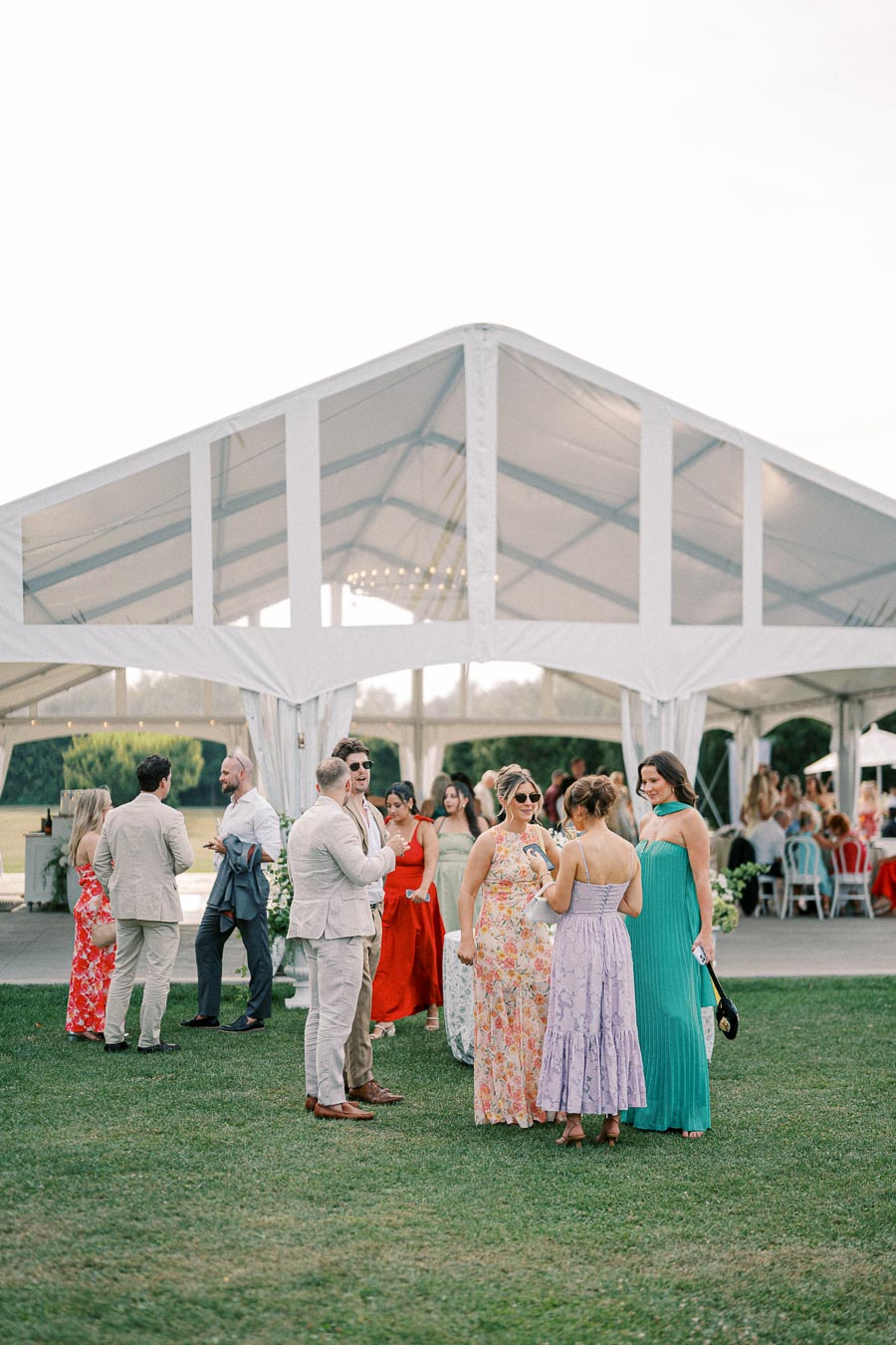 A group of elegantly dressed people enjoys an outdoor event beneath a large white tent on a sunny day.