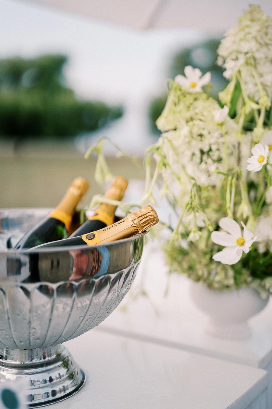 A chilled silver ice bucket holding three bottles of champagne, set on a white table with a vase of white flowers, creating an elegant outdoor setting.