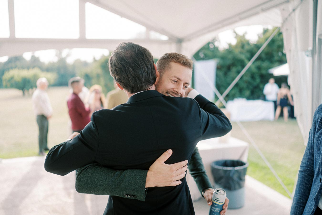 Two men in suits embracing at an outdoor event under a tent, with one holding a can, while guests converse in the background.