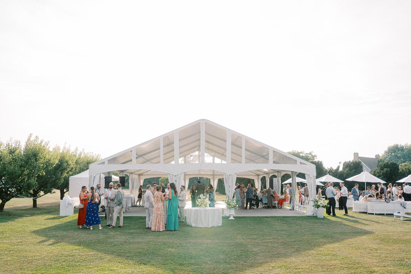 Outdoor wedding reception under a large white marquee tent with guests mingling on a sunny day.