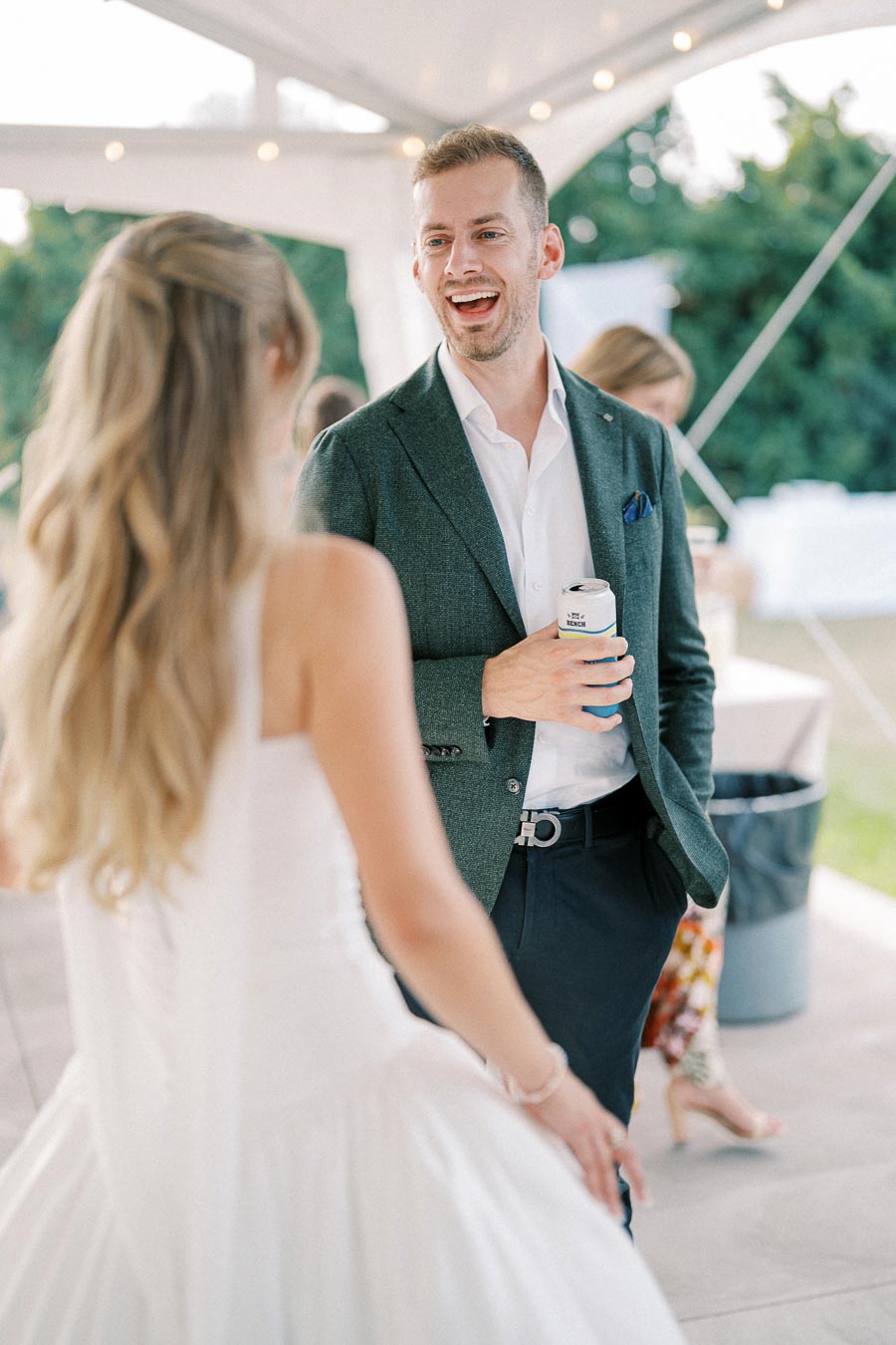 A man in a green blazer and white shirt is laughing and holding a beverage can while talking to a woman in a white dress at an outdoor wedding reception under a tent with string lights.