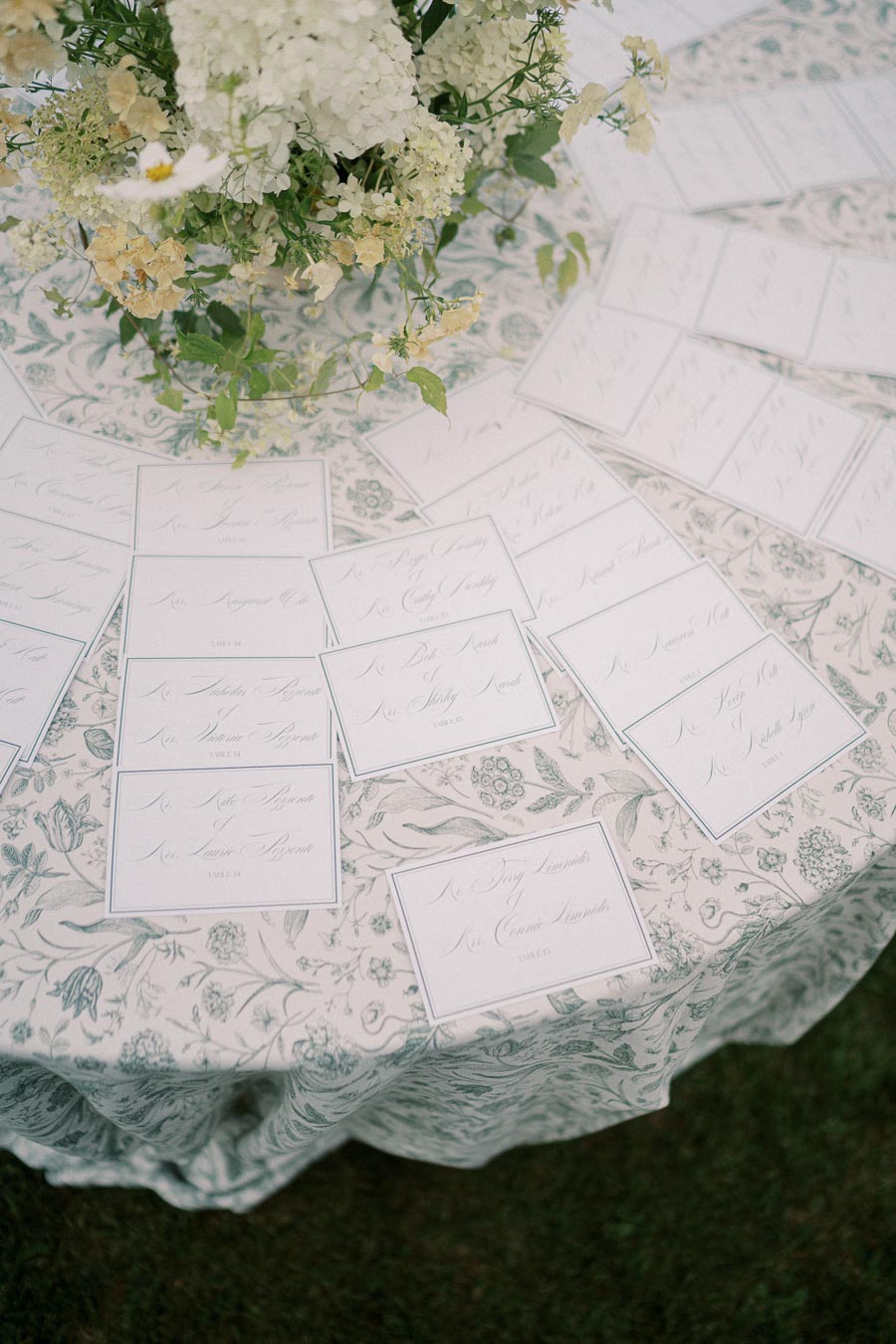 Elegant wedding seating cards with calligraphy on a floral tablecloth, adorned with a centerpiece of lush white and cream flowers.