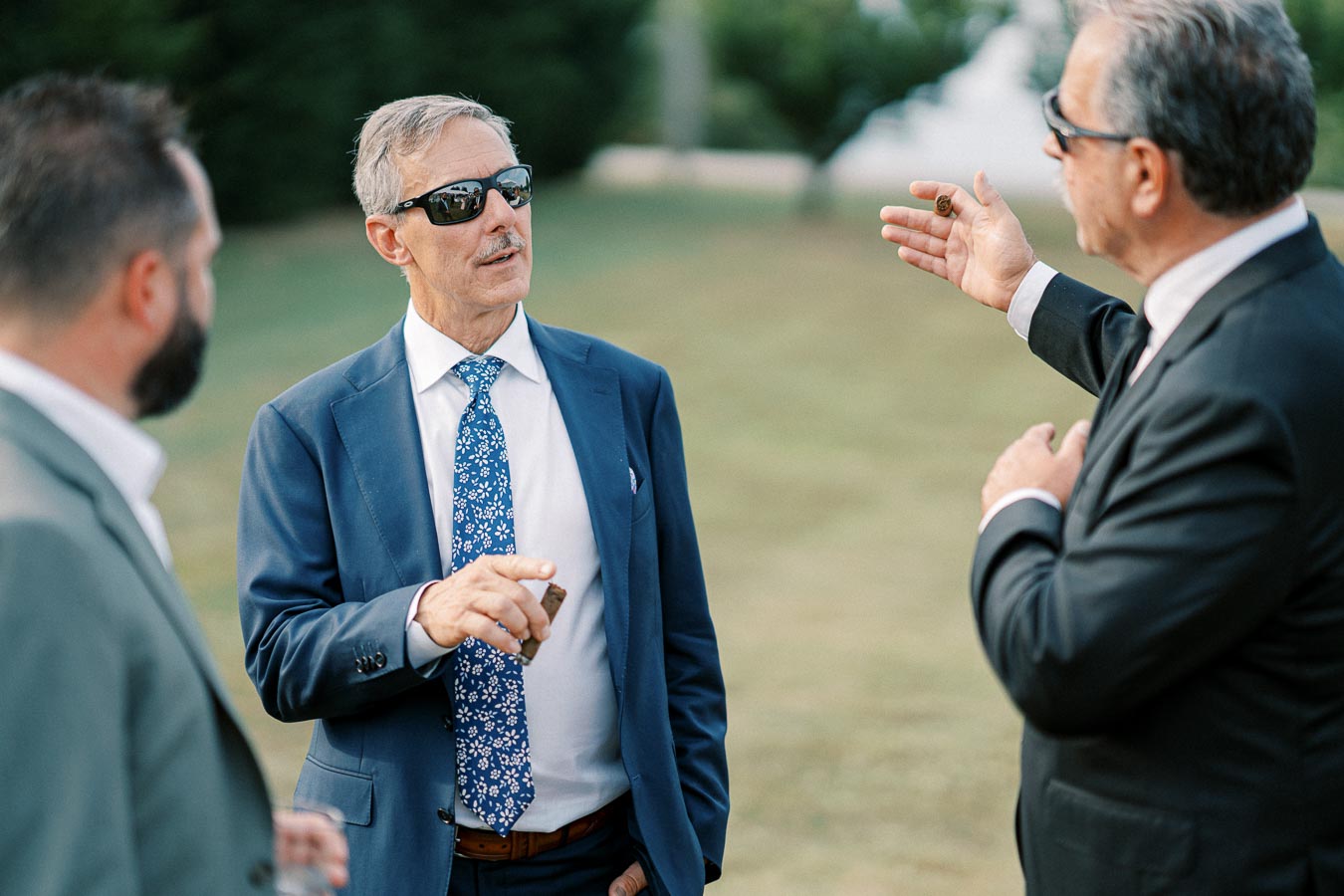 Three men in suits and sunglasses engaged in conversation outdoors, with one holding a cigar, in a professional and relaxed setting.