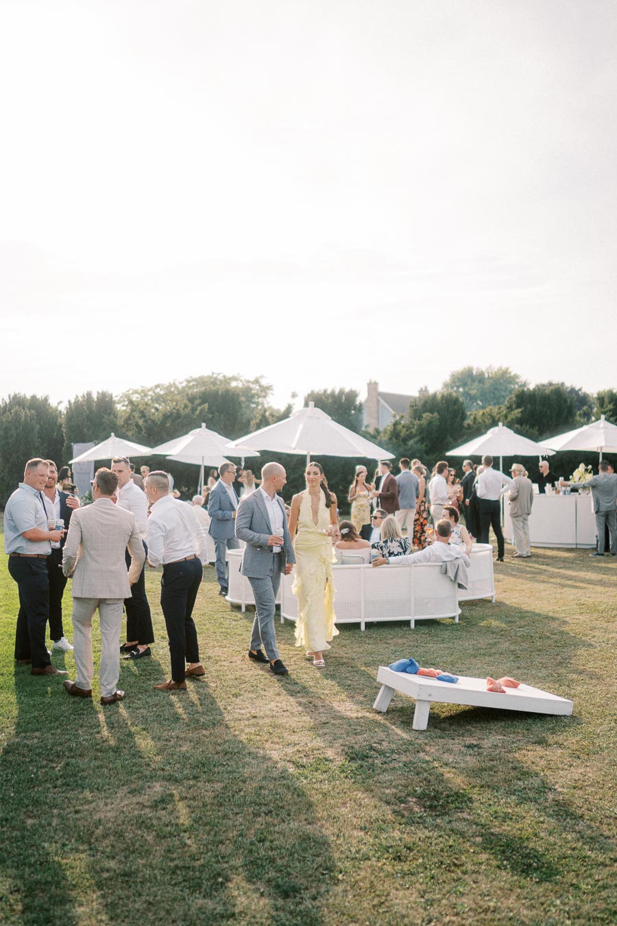 Outdoor wedding reception with elegantly dressed guests socializing under white umbrellas, with a lawn game visible in the foreground.