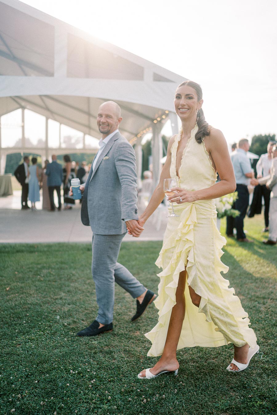 A couple holding hands at an outdoor event, dressed elegantly in a grey suit and a yellow ruffled dress, walking on a lawn with a tent and guests in the background under sunny skies.