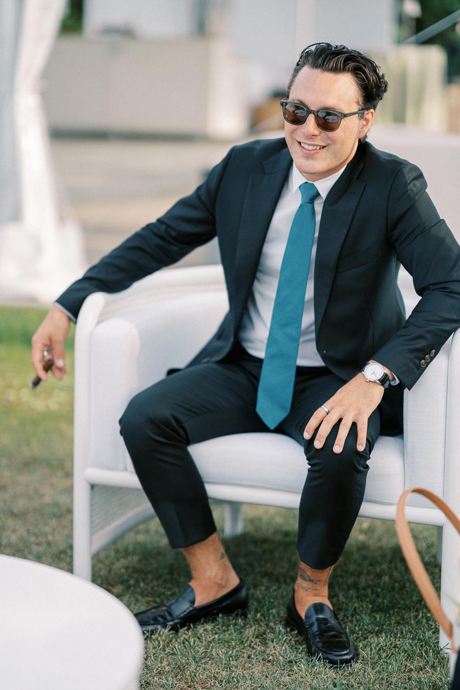 A stylish man in sunglasses, wearing a black suit with a teal tie, sitting casually on a white chair outdoors, exuding confidence and style.