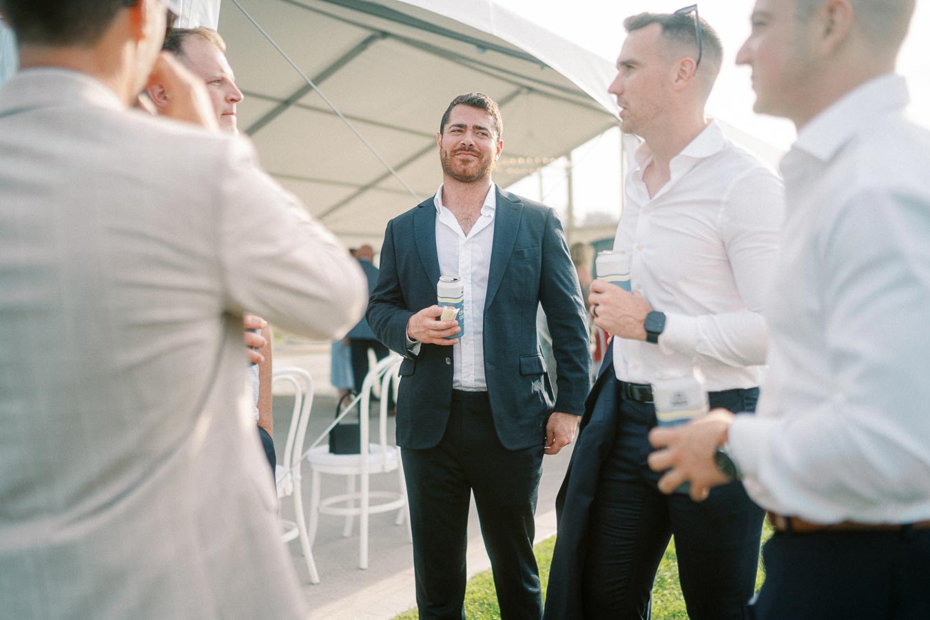 A group of professionally dressed men socializing outdoors at an event, some holding drinks, with a tent and chairs in the background.
