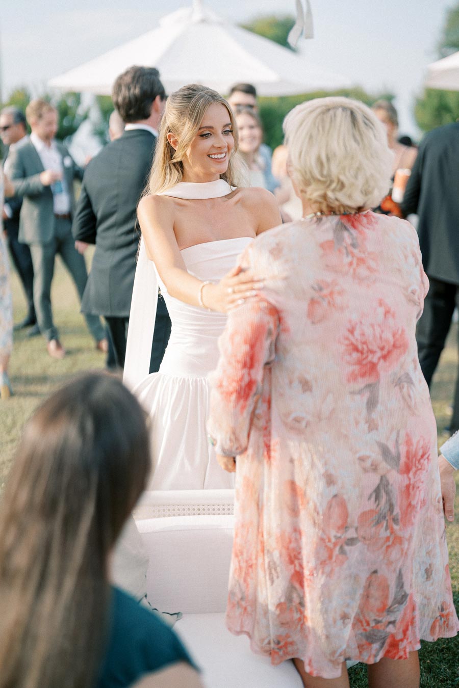 A young woman in an elegant white dress happily greets an older woman in a floral outfit at an outdoor gathering, surrounded by guests in formal attire and sunny weather.