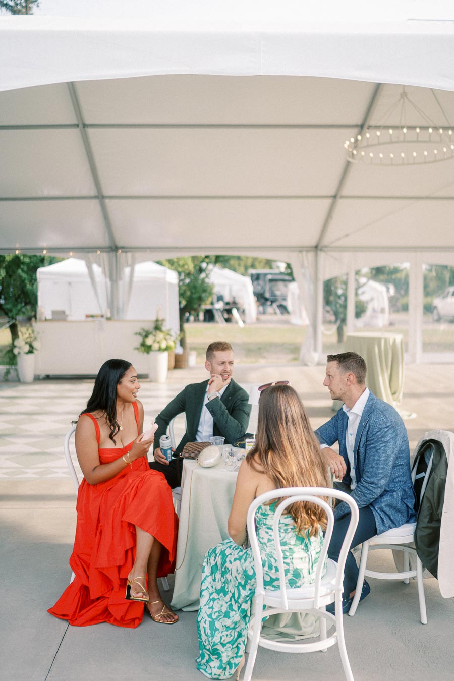 A group of four elegantly dressed people sitting at a table under a large outdoor tent, engaged in conversation during a daytime event.