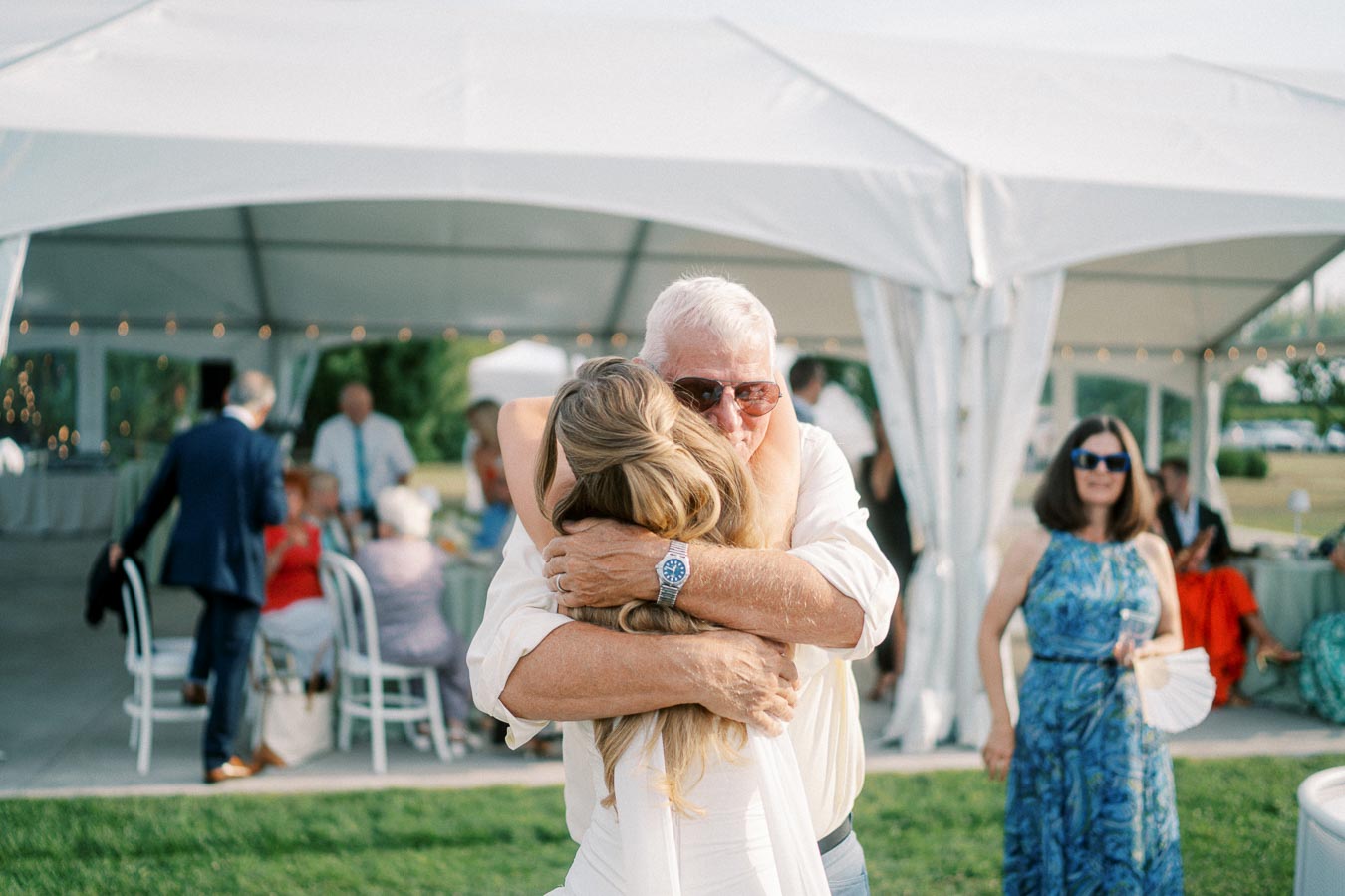 Elderly man embracing a woman warmly under a white event tent at an outdoor gathering, surrounded by well-dressed guests.