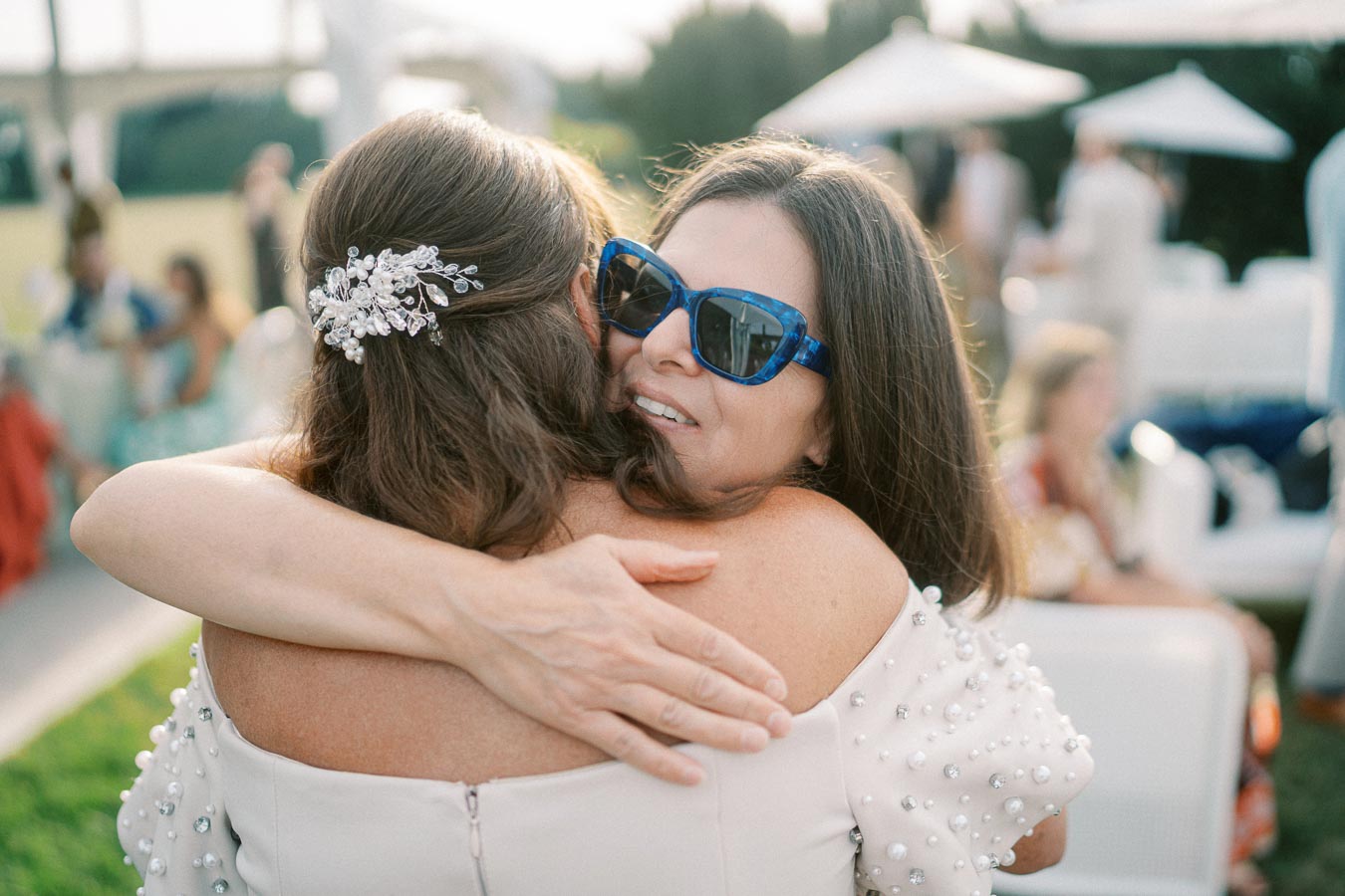 Two women embracing at an outdoor event, one wearing sunglasses and decorative hairpiece.