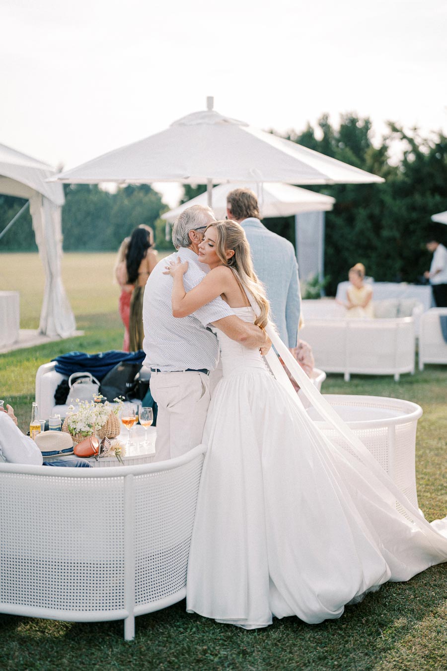 A bride in an elegant white gown embraces an older guest at an outdoor wedding reception, with stylish seating and umbrellas in the background.